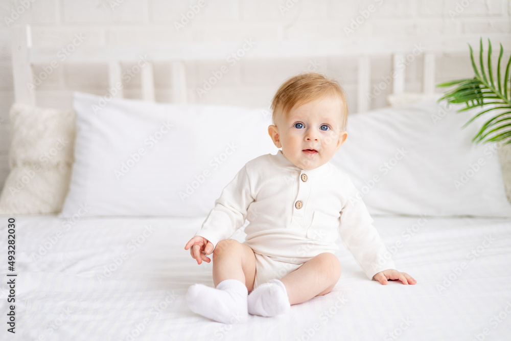 portrait of a 6-month-old blond boy sitting on a large bed in a bright bedroom and smiling in a cotton bodysuit, the concept of children's goods portrait of a 6-month-old blond boy sitting on a large bed in a bright bedroom and smiling in a cotton bodysuit, the concept of children's goods
