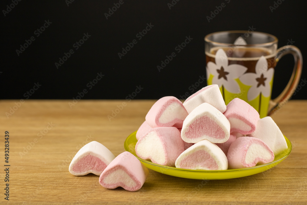 Colored marshmallows sweets in the saucer on around wooden textured background. A cup of aromatic coffee and dessert pink and white in the form of hearts. Cooking concept delicious sweet food. Colored marshmallows sweets in the saucer on around wooden textured background. A cup of aromatic coffee and dessert pink and white in the form of hearts. Cooking concept delicious sweet food.