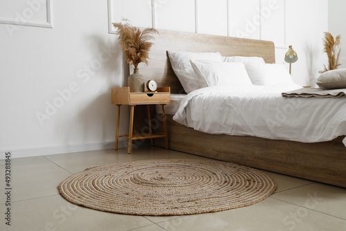 Comfortable bed, wicker rug and nightstand table with dry reeds near white wall