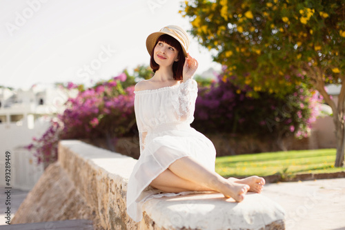 Lovely smiling lady with red hair posing with hand up touching strow hat standing on blur background. Close-up portrait of young woman spending time in tropical town in summer morning.