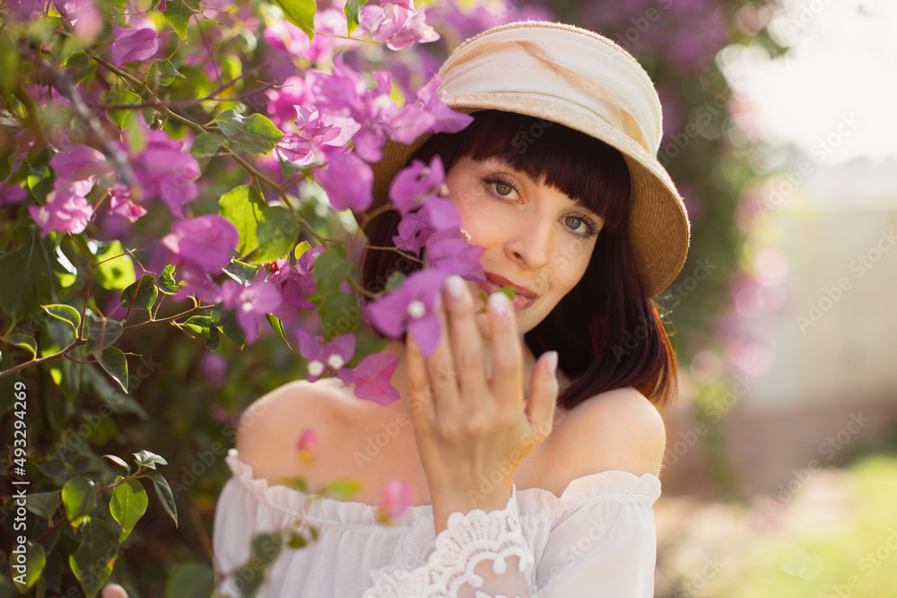 Charming young girl in beautiful flower garden in white dress and straw hat. Red haired girl enjoying the aroma of pink flowers. Woman smelling flowers in summer park with blooming Charming young girl in beautiful flower garden in white dress and straw hat. Red haired girl enjoying the aroma of pink flowers. Woman smelling flowers in summer park with blooming