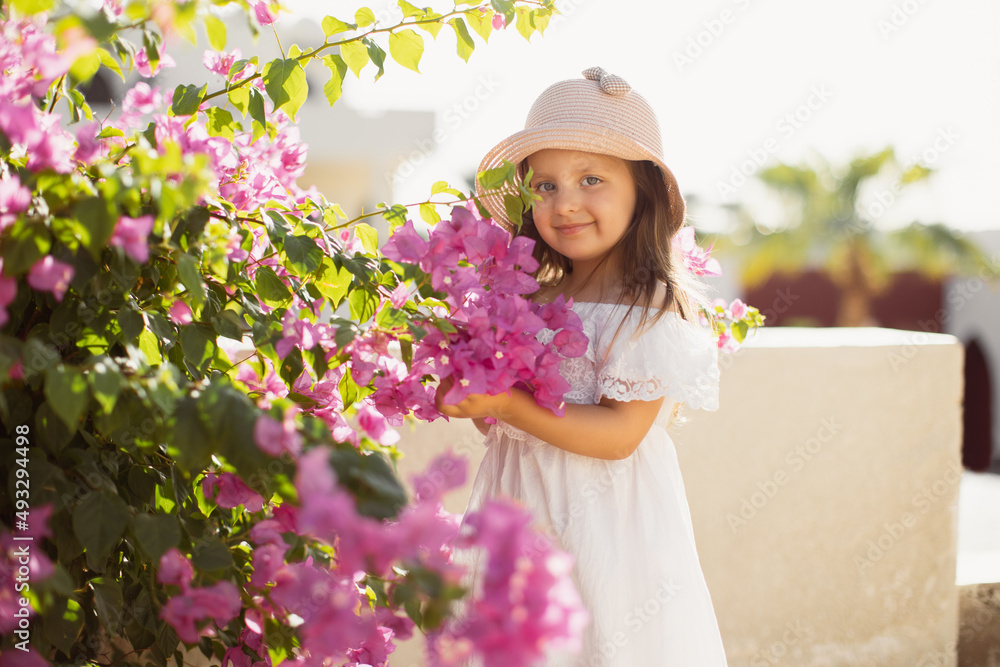 Fototapeta premium Beautiful little cute girl with long blond hair in white dress and straw hat enjoying pink spring blooming. Little preschool girl in near tree with flowers. Springtime.
