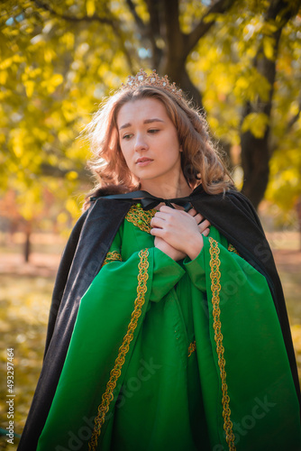 A beautiful girl in a medieval green dress with golden braid. Queen in a cloak and a blio dress in the autumn forest.
