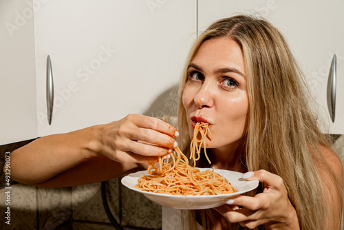 Daring sexy long-legged beauty in a white bodysuit eats spaghetti with tomato paste with her hands. Modern housewife eats pasta in a modern scandinavian kitchen