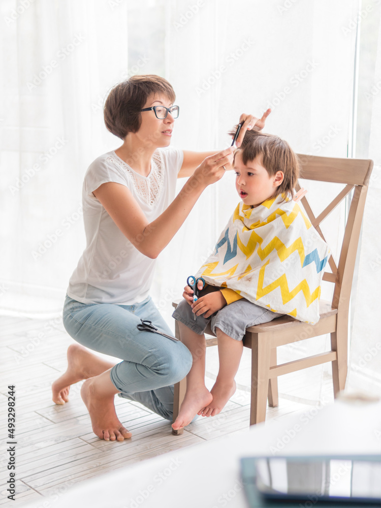 Mother cuts her son's hair by herself. Little boy sits, covered with cloth, and holds pair of scissors. New normal in case of coronavirus COVID-19 quarantine. Beauty at home lifestyle. Mother cuts her son's hair by herself. Little boy sits, covered with cloth, and holds pair of scissors. New normal in case of coronavirus COVID-19 quarantine. Beauty at home lifestyle.
