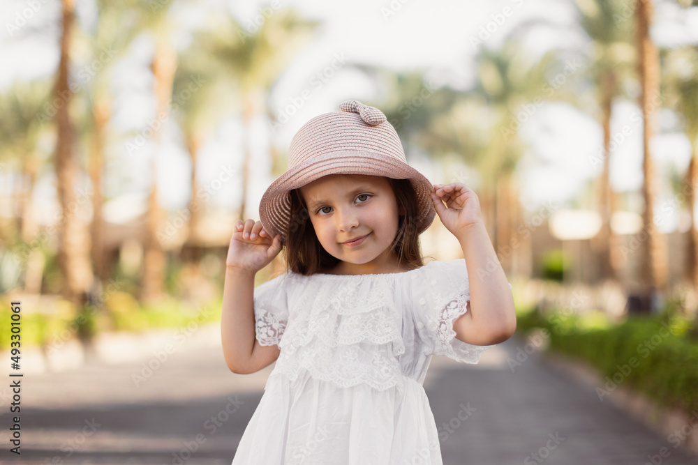 Close up portrait of a happy little girl in white boho dress and straw hat on the background of palm trees. Summer vacations concept.