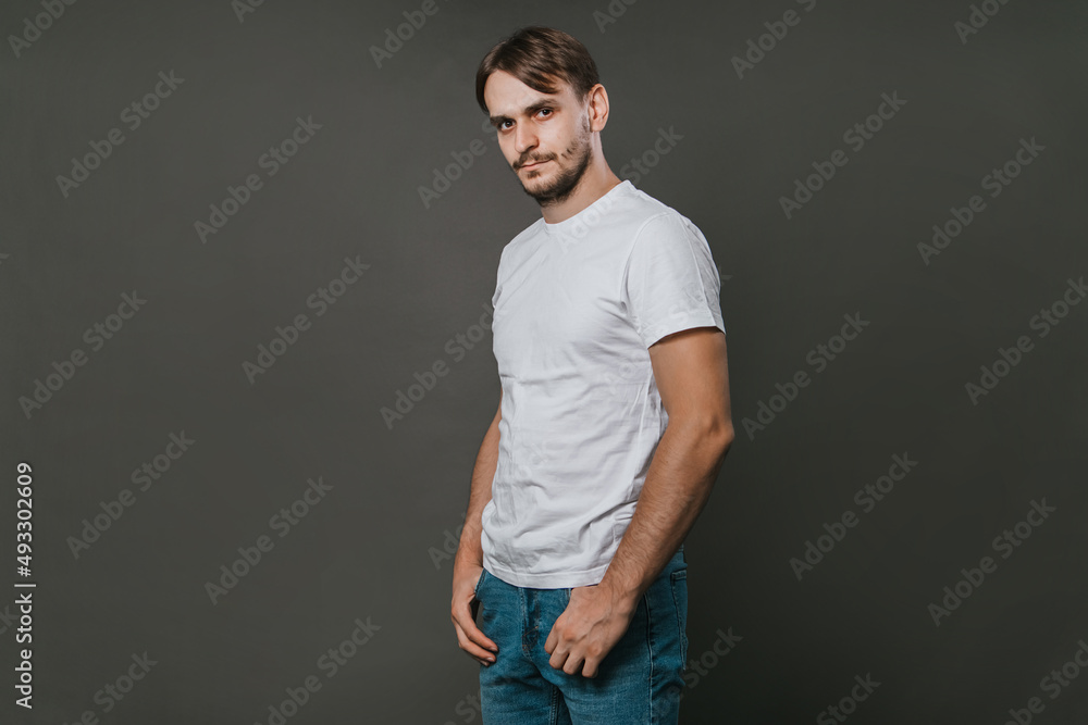 A handsome man in a white t-shirt and jeans stands on a gray background. Studio photo.
