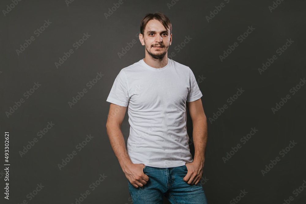 A handsome man in a white t-shirt and jeans stands on a gray background. Studio photo. A handsome man in a white t-shirt and jeans stands on a gray background. Studio photo.