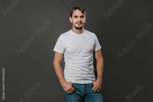 A handsome man in a white t-shirt and jeans stands on a gray background. Studio photo.