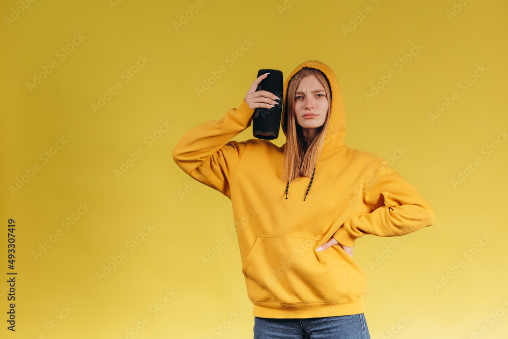 Funny young girl looking, waving hand, holding portable wireless bluetooth music speaker, isolated on yellow background. Girl in a yellow hoodie Funny young girl looking, waving hand, holding portable wireless bluetooth music speaker, isolated on yellow background. Girl in a yellow hoodie