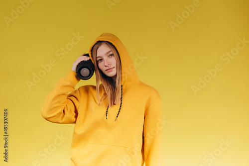 Funny young girl looking, waving hand, holding portable wireless bluetooth music speaker, isolated on yellow background. Girl in a yellow hoodie photo