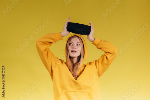 Funny young girl looking, waving hand, holding portable wireless bluetooth music speaker, isolated on yellow background. Girl in a yellow hoodie © Igor