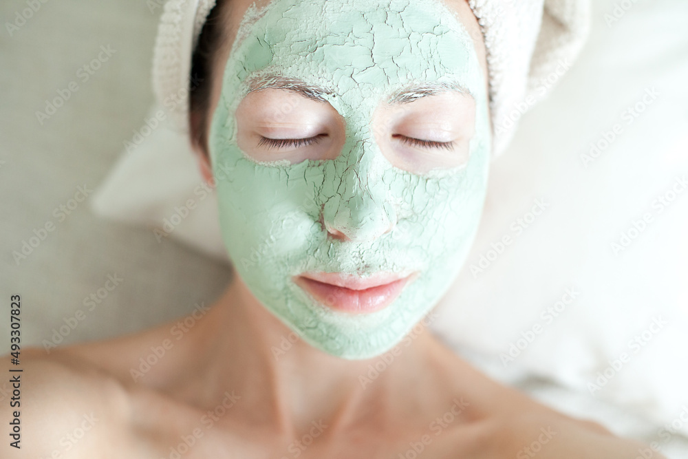 Fototapeta premium Young woman with green clay cosmetic mask on her face - skincare and spa treatment beauty routine, relaxing moments