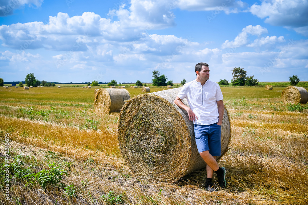 Happy farmer is standing beside bales of hay. He is examining straw after successful harvesting. Strong handsome man portrait on agricultural field Happy farmer is standing beside bales of hay. He is examining straw after successful harvesting. Strong handsome man portrait on agricultural field