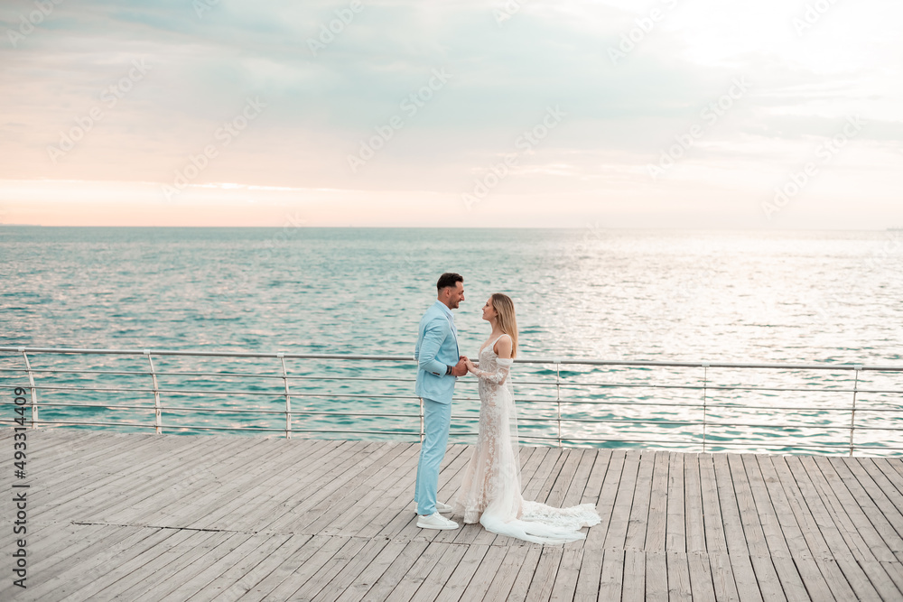 Young bride, fashionable, blonde model in luxury wedding dress with groom on the coast. Couple walks at the pier on sunrise