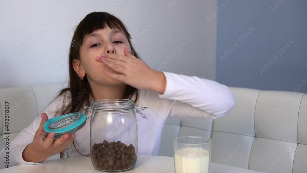 Little girl eats chocolate corn balls from glass jar sitting at white table. Child have breakfast Little girl eats chocolate corn balls from glass jar sitting at white table. Child have breakfast