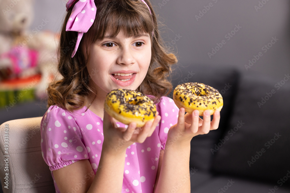 Naklejka premium Cheerful school girl playing with cakes indoors. Funny teenager girl having fun with colorful donuts at modern home. Portrait of sweet girl choosing between two donuts in home room.