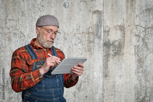 confident senior man wearing glasses, a flannel shirt and overalls is making notes or sketches on a digital tablet photo