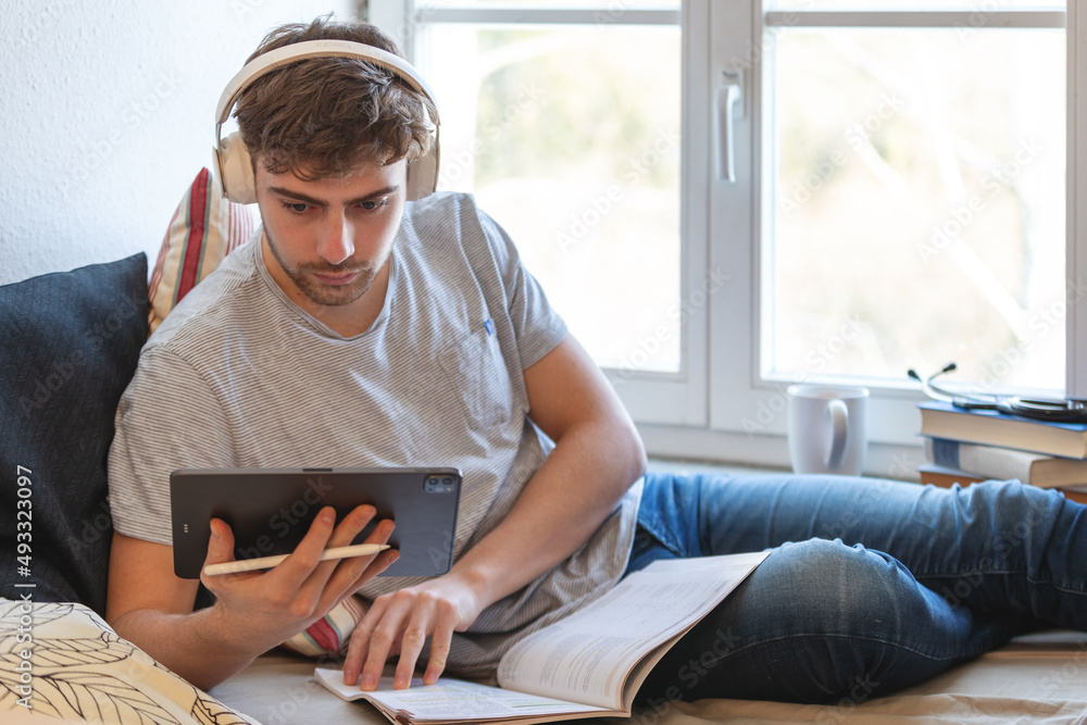 Junger Medizinstudent studiert online von zu Hause aus. Er liegt auf seinem Bett vor einem Fenster, trägt Kopfhörer, blickt konzentriert in sein Tablet und blättert in Unterlagen