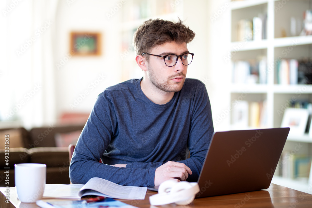 Junger hutaussehender Mann mit Brille und Bart sitzt am Schreibtisch mit einem Laptop und denkt nach. Auf dem Tisch steht eine Tassen fliegen Kopfhörer. Junger hutaussehender Mann mit Brille und Bart sitzt am Schreibtisch mit einem Laptop und denkt nach. Auf dem Tisch steht eine Tassen fliegen Kopfhörer.