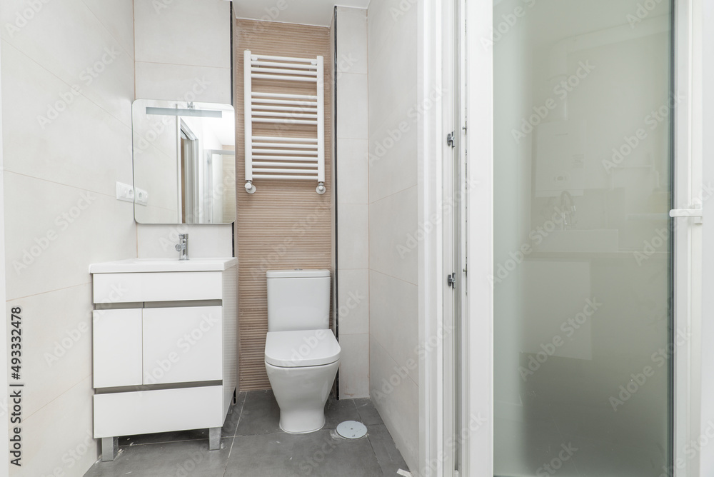 Tiled toilet with large white tiles, white porcelain sink with glass mirror and white wooden cabinet and white aluminum towel radiator