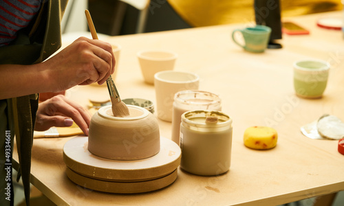 Close-up of girl painting clay mug with glaze. Woman coloring pottery in workshop with a paintbrush. Painter in green apron glazing clay pot.