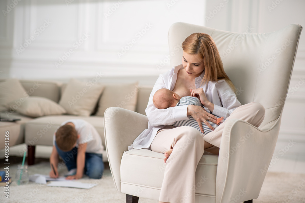 Mom breastfeeds baby sitting on a chair. Behind the eldest son draws a picture for mom and sister. Mom breastfeeds baby sitting on a chair. Behind the eldest son draws a picture for mom and sister.