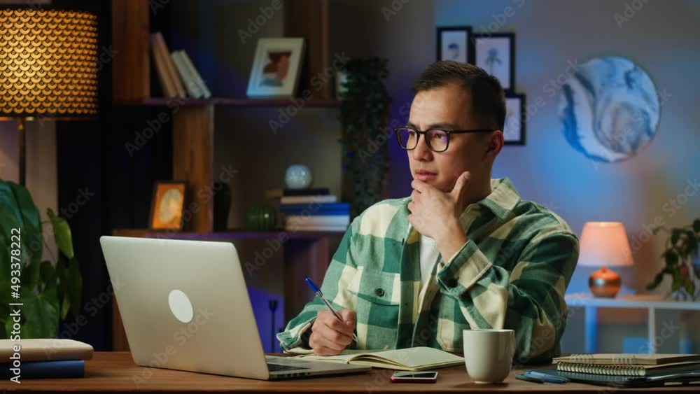 Asian man typing on laptop and writing notes, sitting in living room. Korean young guy working remotely at home during coronavirus lockdown. Businessman using computer for remote work, freelance. Asian man typing on laptop and writing notes, sitting in living room. Korean young guy working remotely at home during coronavirus lockdown. Businessman using computer for remote work, freelance.