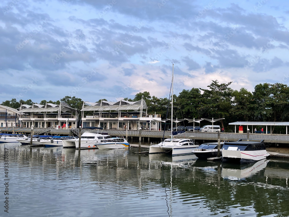 Boat station, view from the water. People and cars on the embankment. Motor yachts and sailing yachts are moored at the pier. Sea transport. Buildings on a coast. Boats, sailboats, cutters for cruise.