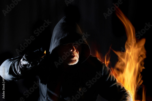 Fire and flames surround the portrait of an arsonist. He is wearing a hooded shirt. The background is black.