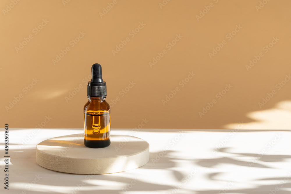 Unmarked amber glass bottle with dropper lid on beige background with floral shadow. Mock-up of facial serum in rays of sunlight. Close-up, copy space