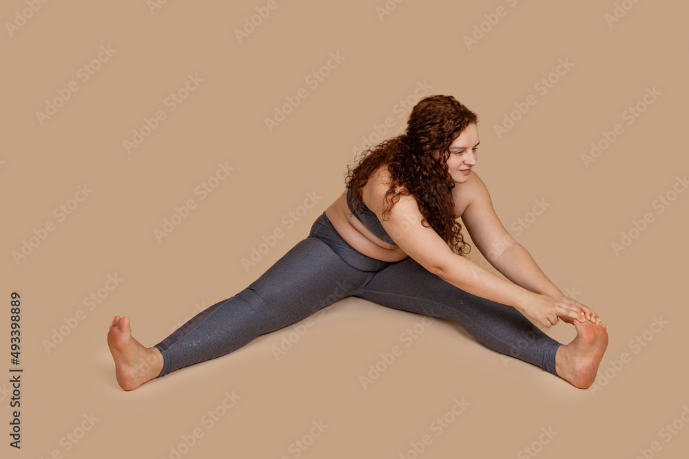 Overweight smiling enjoying woman, with obesity, excess fat in gray sportswear. Tummy, belly flabs incline to leg. Weight loss. Self challenge goal to dieting, body sculpting. Beige background
