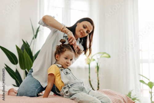 A happy mom combs her little daughter's hair while sitting in bed in the morning