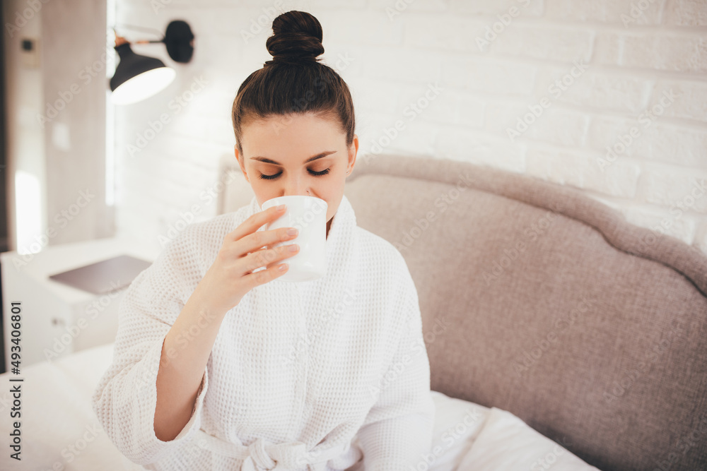 Young woman wearing white bathrobe sitting on bed, drinking hot coffee in her bedroom. Young woman wearing white bathrobe sitting on bed, drinking hot coffee in her bedroom.