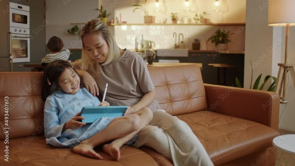 Asian Family With a Daughter do Homework by Using Tablet With Mother Help, Touch Screen Together While Sitting in the Living Room at Home Morning. Concept of Online Learning at Home. Asian Family With a Daughter do Homework by Using Tablet With Mother Help, Touch Screen Together While Sitting in the Living Room at Home Morning. Concept of Online Learning at Home.