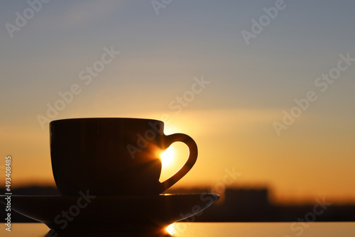 Silhouette of coffee or tea cup on background of sunset sky and shining sun. View from the window to evening city