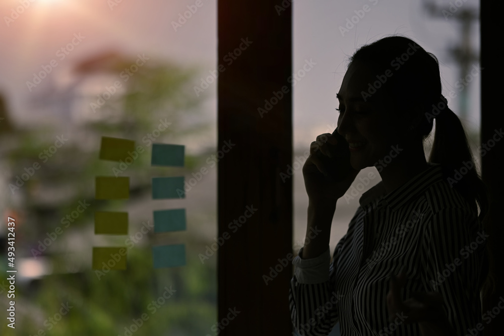 Silhouette of young businesswoman talking on mobile phone while standing near glass window with view on sunset in city.