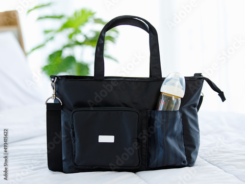 Closeup studio shot of black color multifunction multipurpose utility newborn toddler accessories handbag with milk baby bottle and plastic cap in elastic pocket with handle and strap placed on bed photo