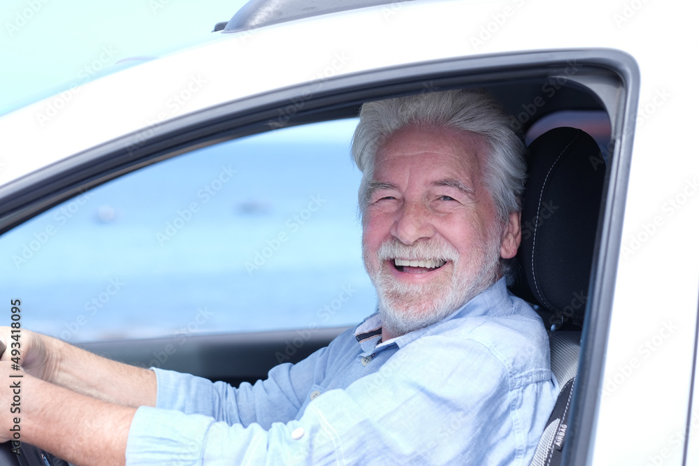Beautiful smiling adult white-haired senior man sitting inside the car touching the steering wheel looking at camera. Elderly grandparent with white beard enjoying travel. Horizon over water Beautiful smiling adult white-haired senior man sitting inside the car touching the steering wheel looking at camera. Elderly grandparent with white beard enjoying travel. Horizon over water