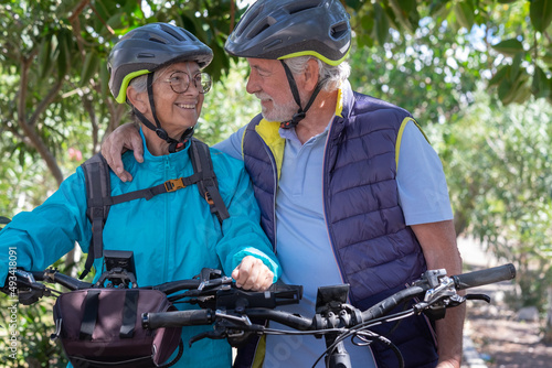 Active senior caucasian couple with electrobikes standing outdoors in the park. Two happy elderly people looking in the eyes enjoying nature and healthy lifestyle