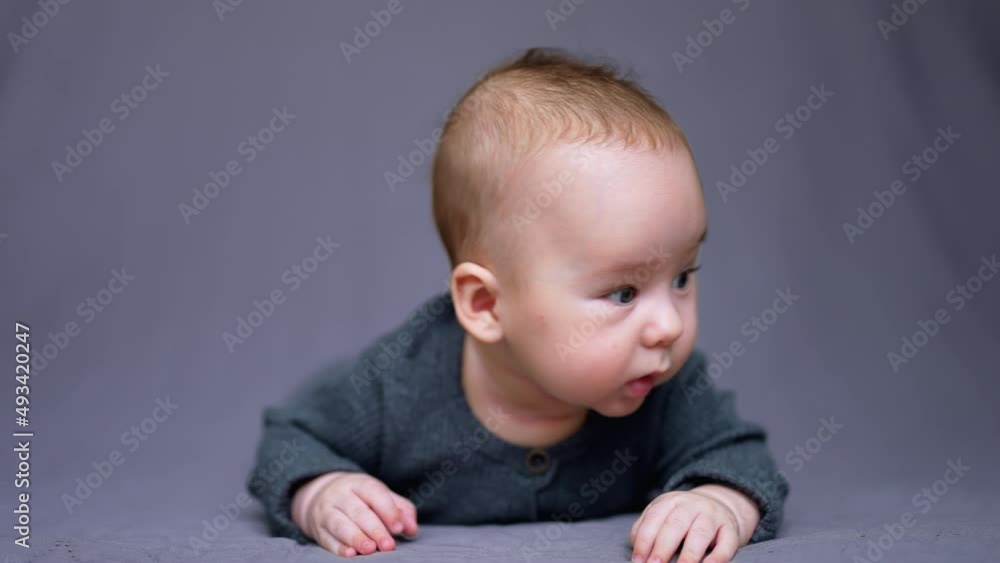 Adorable little child in grey clothes lies on belly in front of camera. Sweet funny boy with plump cheeks making faces. Close up. Adorable little child in grey clothes lies on belly in front of camera. Sweet funny boy with plump cheeks making faces. Close up.