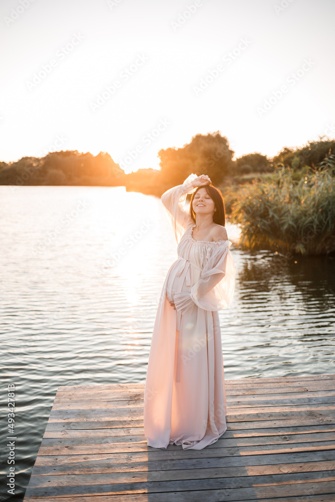 A young pregnant woman in a chiffon dress stands on a pier by the river against the backdrop of an orange sunset