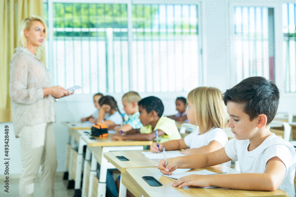 Focused preteen pupils studying in classroom with female teacher, writing exercises in workbooks