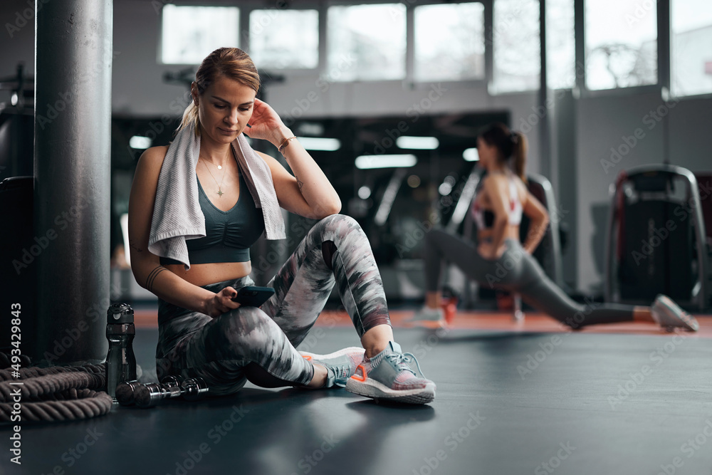Pretty woman working out in a gym, making pause and using smartphone. Adult pretty sporty lady with beautiful shaped body.