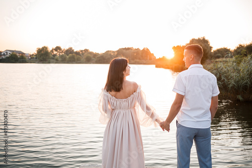 A pregnant woman and her husband stand on the pier and look at each other, a beautiful river and sunset, future parents spend time outdoors, a beautiful young mother