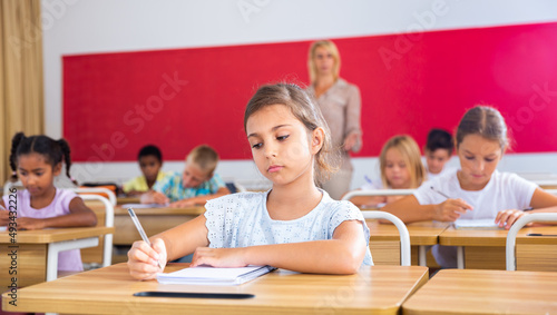 Portrait of focused tween schoolgirl writing exercises in workbook in classroom during lesson