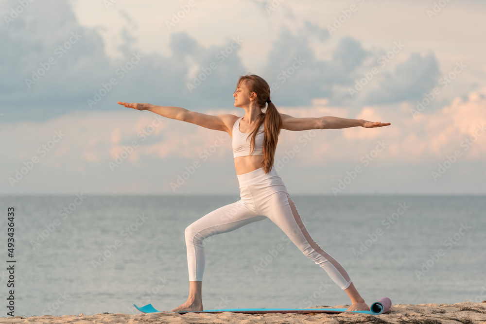Young woman in the warrior pose outdoors. Girl practices yoga at dawn on sea background. Yoga at the beach. Young woman in the warrior pose outdoors. Girl practices yoga at dawn on sea background. Yoga at the beach.