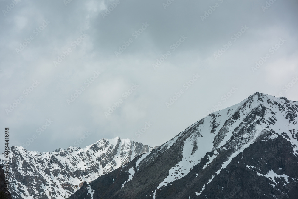 Awesome landscape with high snowy mountain range with sharp rocks in sunlight in cloudy sky. Dramatic view to snow mountains in changeable weather. Atmospheric scenery with white snow on black rocks.