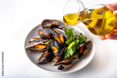 Cooked mussels with parsley leaves in white dish topped with extra virgin olive oil poured from glass decanter on white background