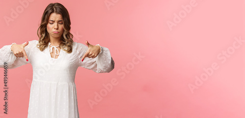 Gloomy, jealous and upset cute blond european woman in white trendy spring dress, sulking as pointing and looking down, see something expensive, cant efford it, strand pink background photo